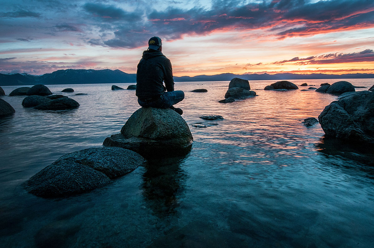 Mindfulness spiritual practice, a young man sitting on a rock by a calm sea or lake at sunset.