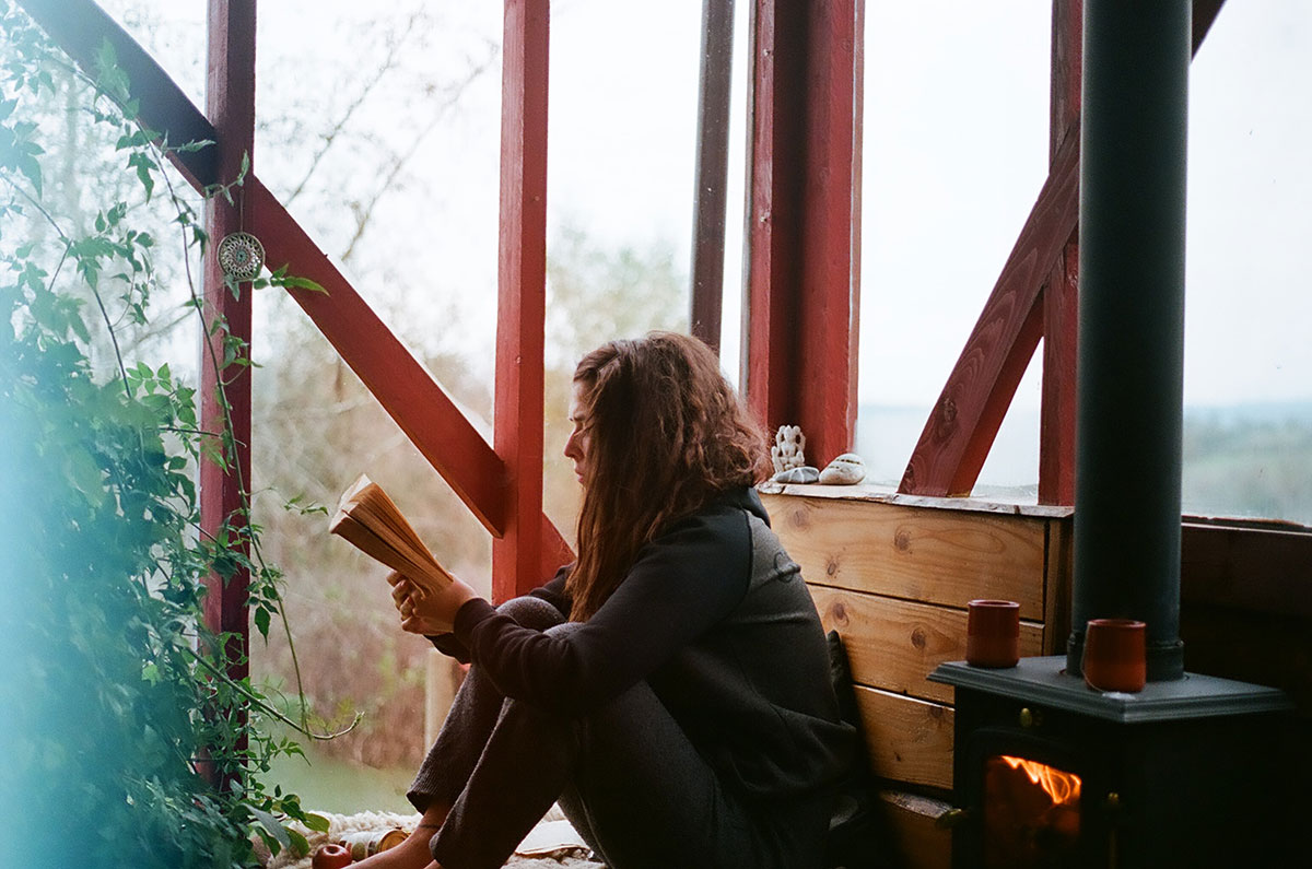 A girl reading a book in a wooden cabin by a wood burning stove.