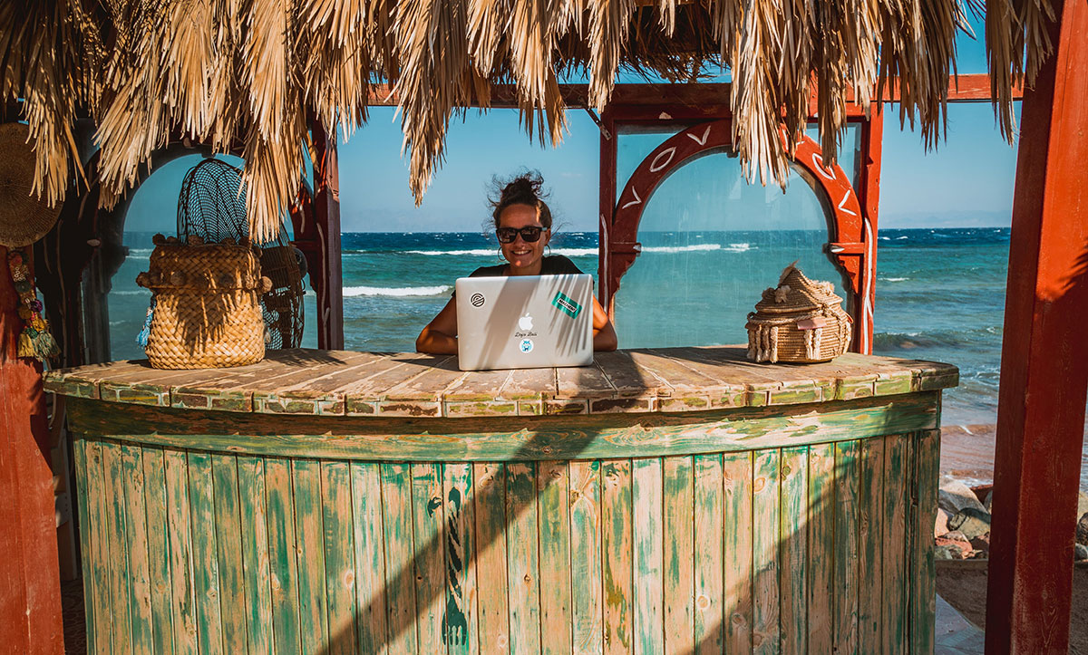 Girl with smile working with laptop in beach bar with sea behind.