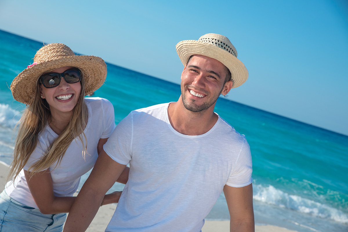 Happy couple in the beach wearing white tops and straw hats.